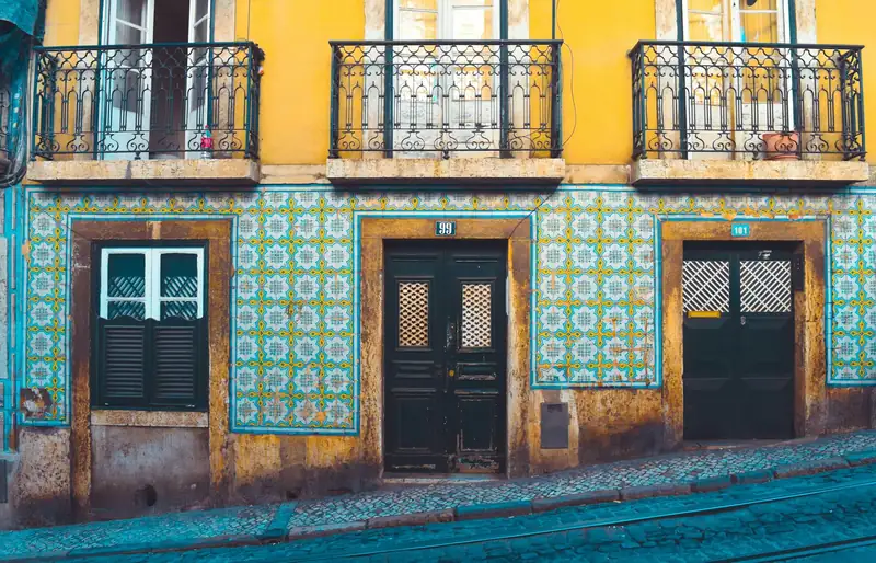 Fachada amarela com azulejos azuis, três varandas de ferro e portas de madeira numa rua empedrada de Lisboa